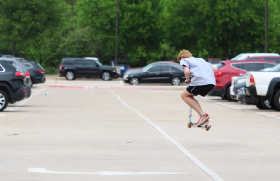 A student does a jump on his scooter in the student parking lot as students are released after fourth period for eighth period release. In the student parking lot students can be seen walking to or from their cars listening to music, talking to their friends and looking at their phones all while dodging traffic. 