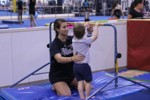 Coppell High School senior Sophia Olson coaches one of her 2 year old students to do a backflip on a bar during Gymnastics practice. Olson helps by supporting the students back to help guide him to flip over. 