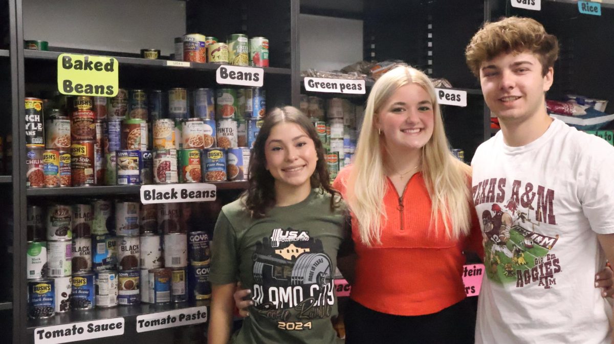 Coppell High School senior Jakie Fuerte, senior Quade Dickert and junior Annabella Perk stand beside the CHS food pantry in room D108 on Dec. 4. The pantry collects donated food to combat food insecurity and support the Coppell community. 